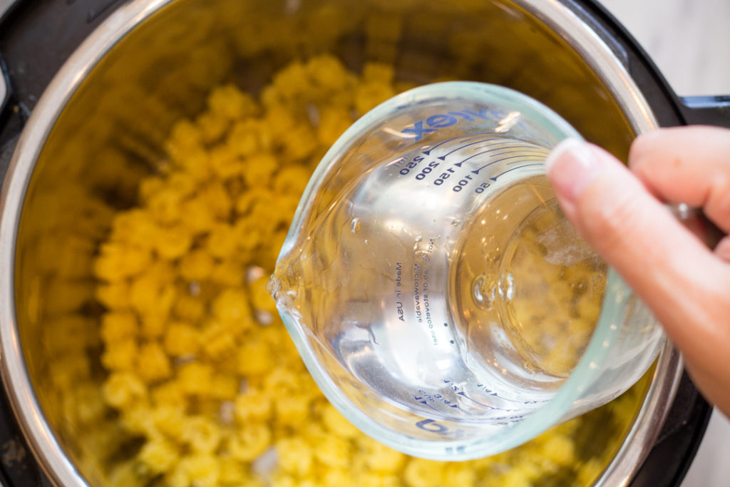 Top view of an instant pot with pasta in it, and a jug of water being poured into it.