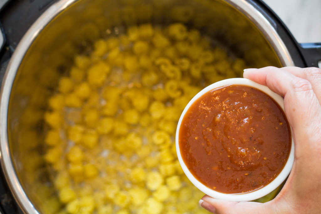 Top view of an instant pot with pasta and water in it, with tomato salsa in a small bowl being held over it.
