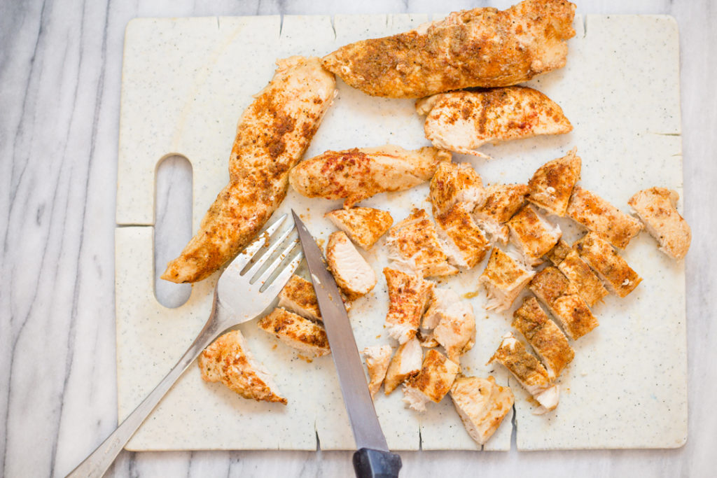 Top view of chicken tenders being cut into cubes with a sharp knife on a white chopping board.