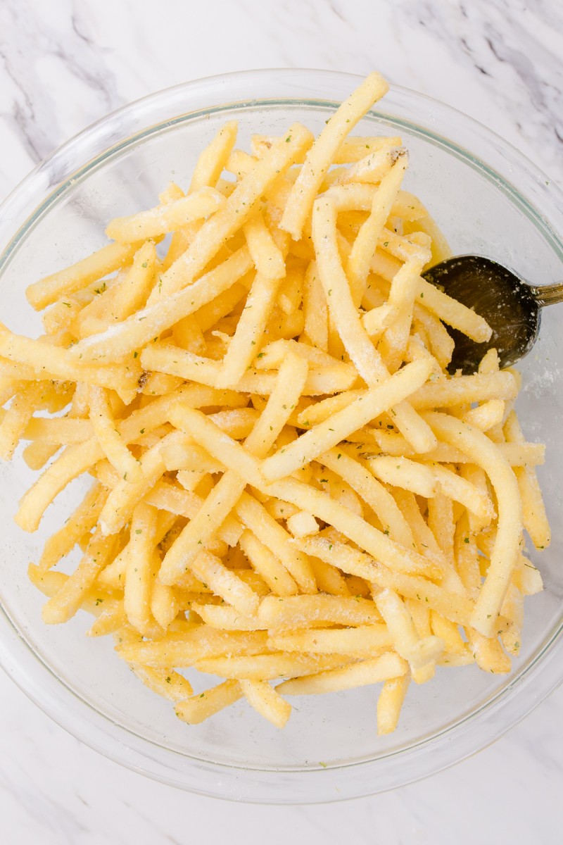 Top view of a glass mixing bowl with fries in it coated with seasonings.