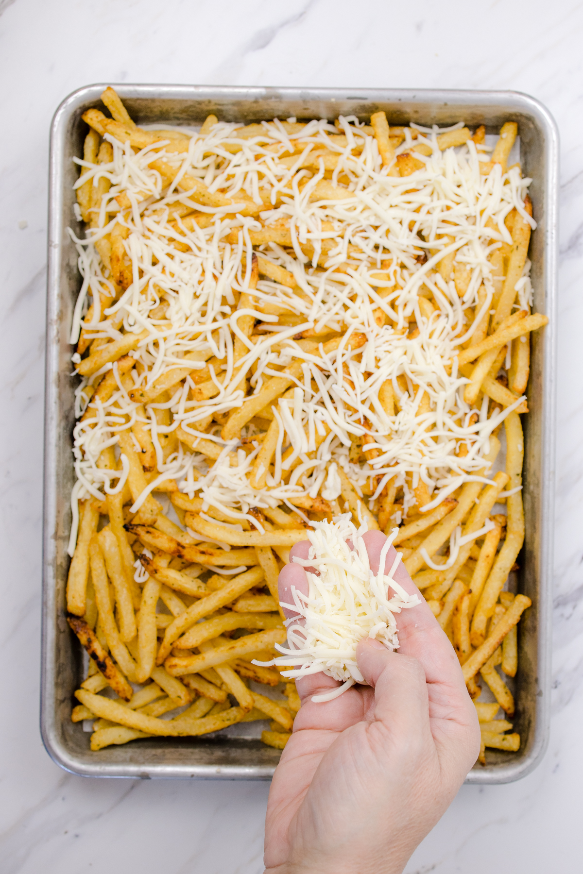 Top view of a baking tray with fries on it being topped with cheese by a hand. 