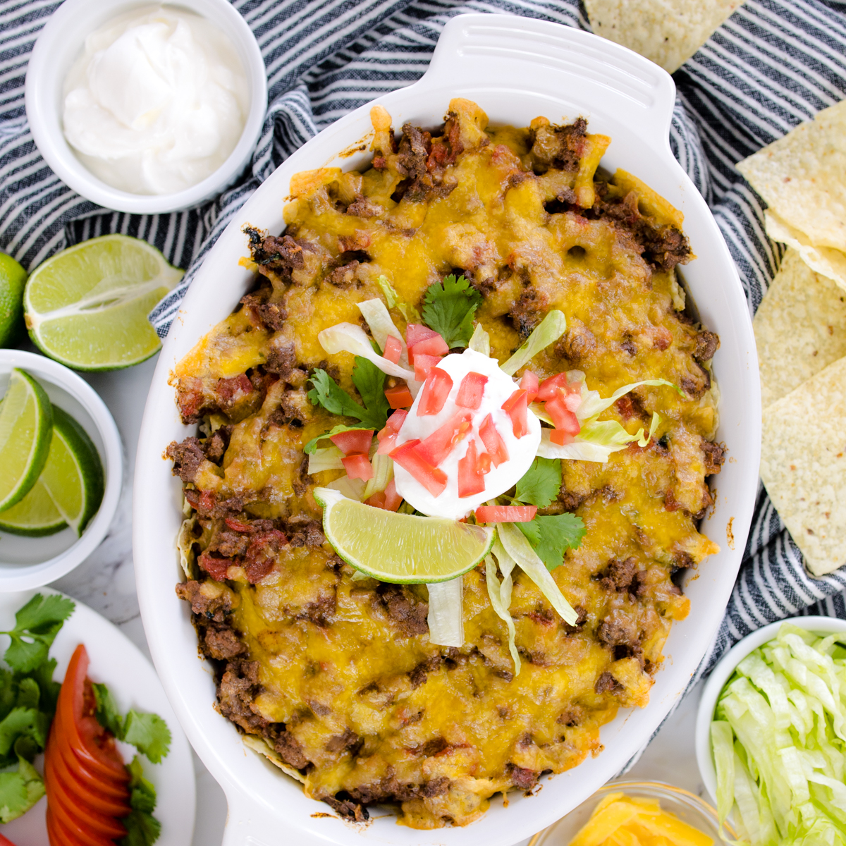 Top view of freshly baked taco caserole with a dollop of sour cream on top of it with some chopped tomato, fresh cilantro, and a lime wedge on top, surrounded by small bowls with cheese, guacamole, and tortilla chips in them.