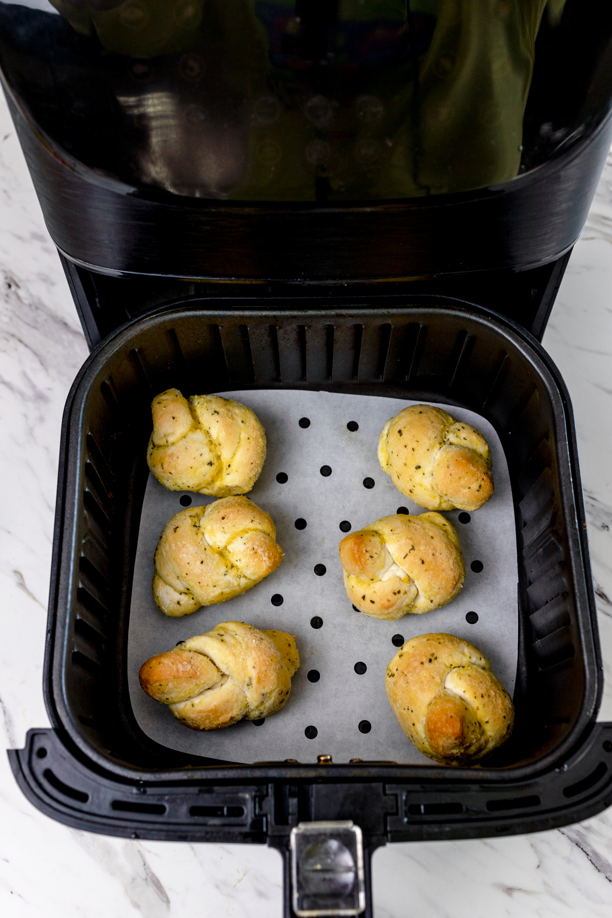 Top view of an air fryer basket pulled out from the air fryer with parchment paper lined on the bottom and frozen garlic knots placed in a single layer on the bottom.
