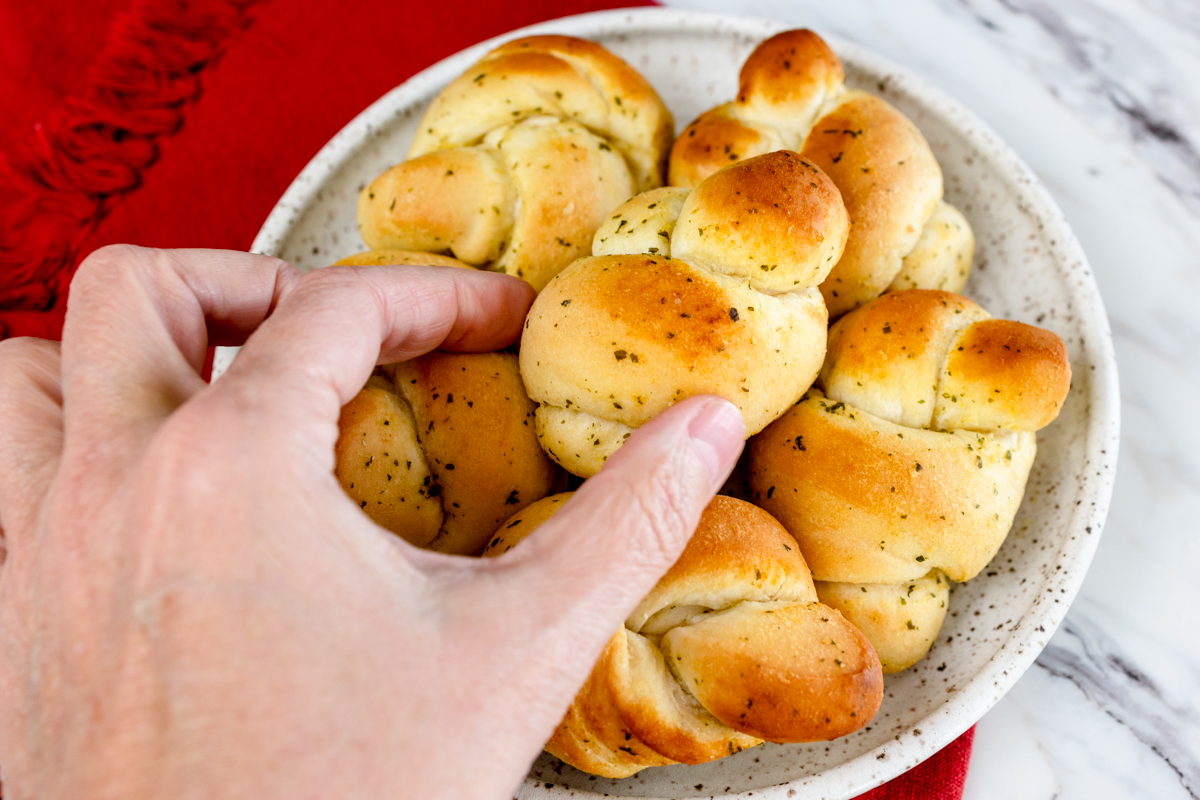 Top view of garlic knots on a white plate, one being lifted up by a hand.