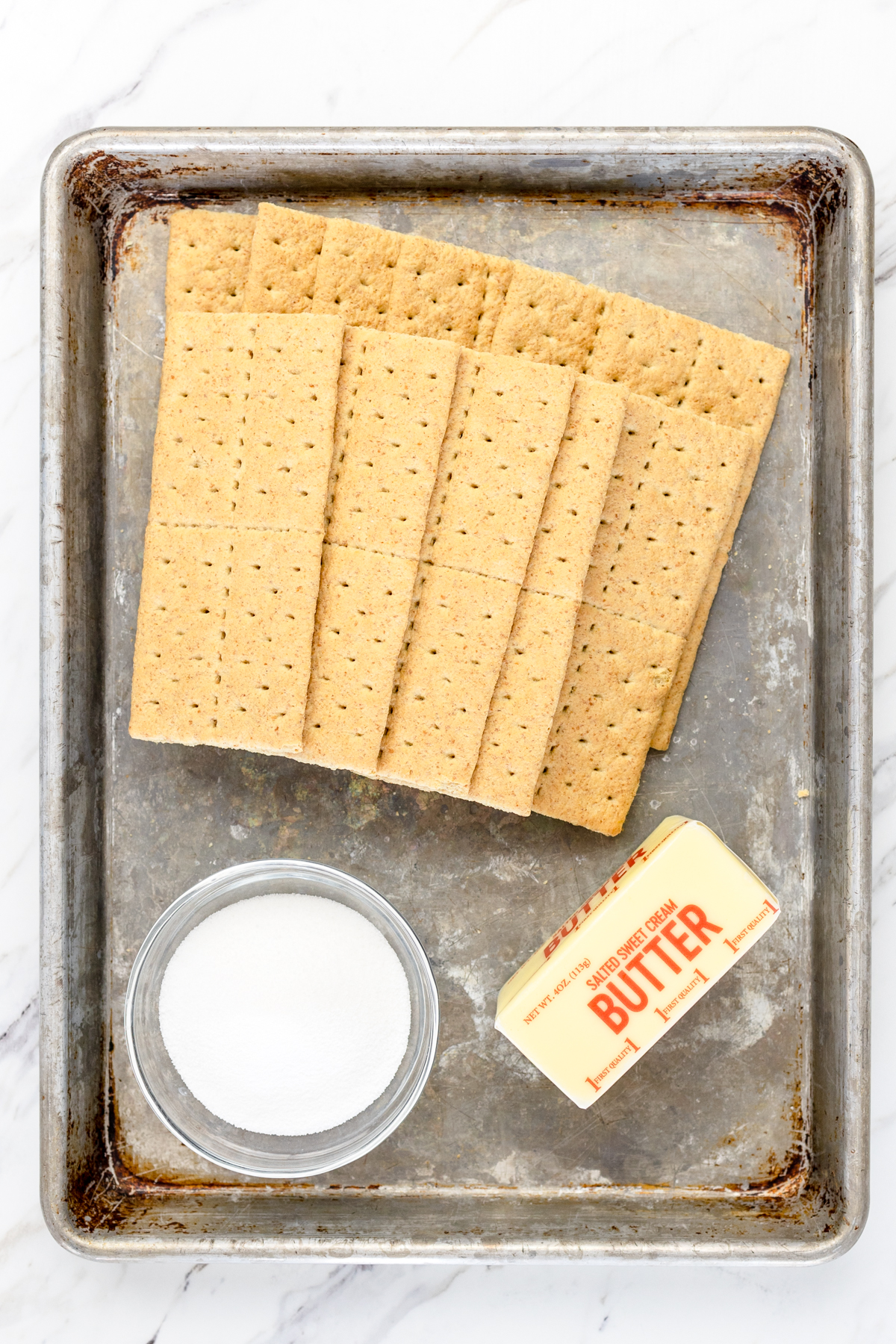 Top view of a baking tray with graham crackers, sugar, and butter on it as the ingredients for a Graham Cracker Crust. 