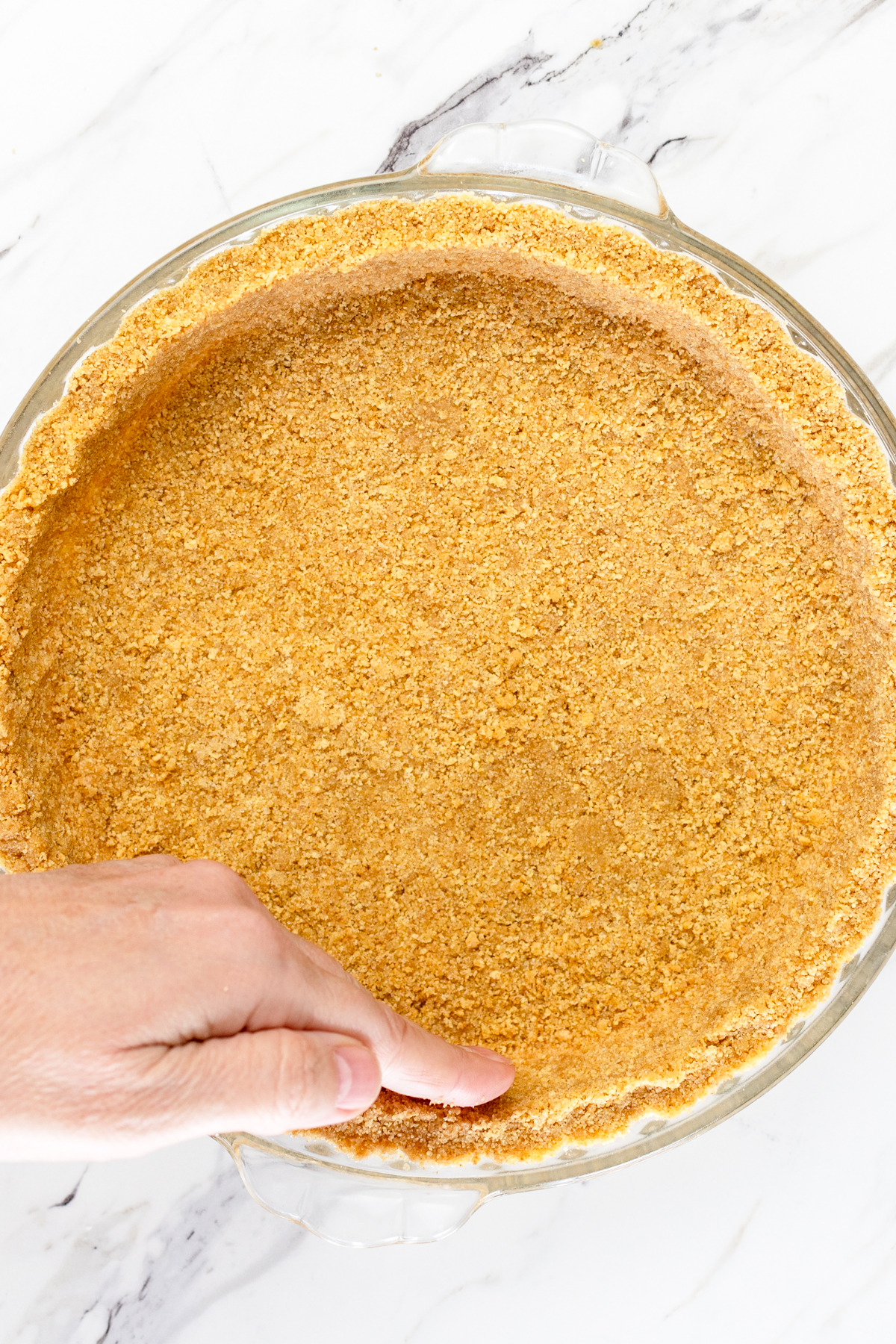 Top view of a glass pie dish with crumbled graham crackers miced with butter and sugar being pressed into the bottom and sides to form a pie crust. 