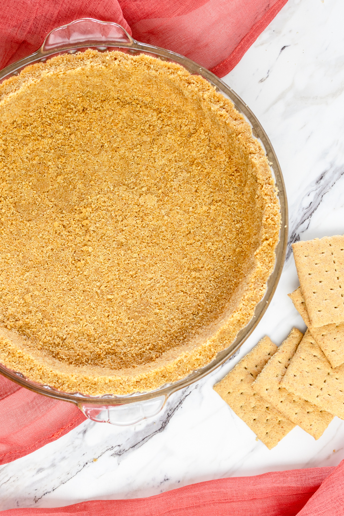 Top view of a graham cracker crust in a glass pie dish with graham crackers lying on the table next to it. 