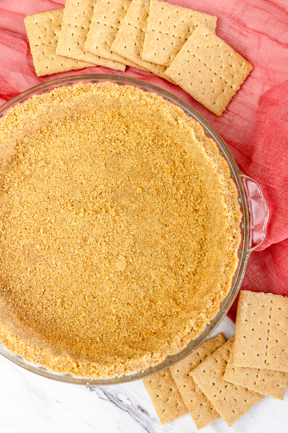 Top view of Graham Cracker crust in a pie dish on a table with a red tea towel, with whole graham crackers next to it, fanned out. 