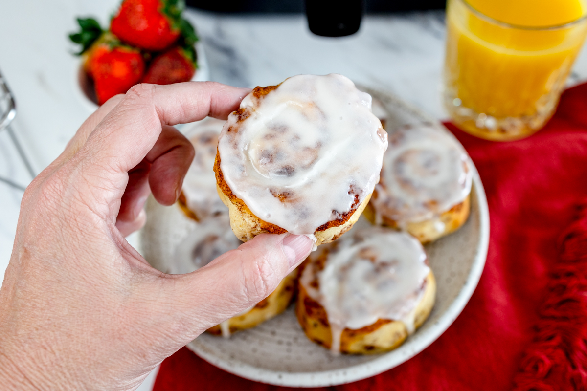 Top view of a glazed cinnamon roll being lifted up by hand off a plate filled with cinnamon rolls. 