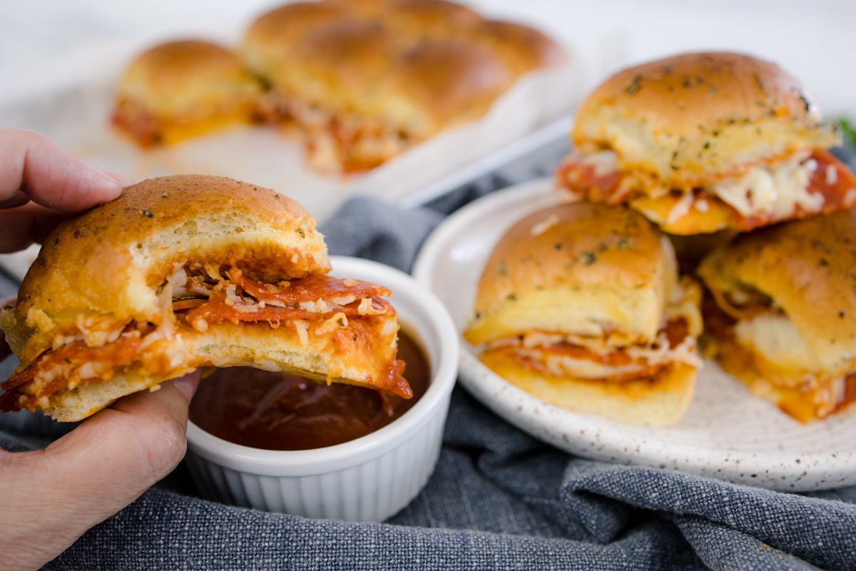 Close up of a pizza slider with a bite taken out of it being dipped into marinara sauce, with more pizza sliders in the background. 