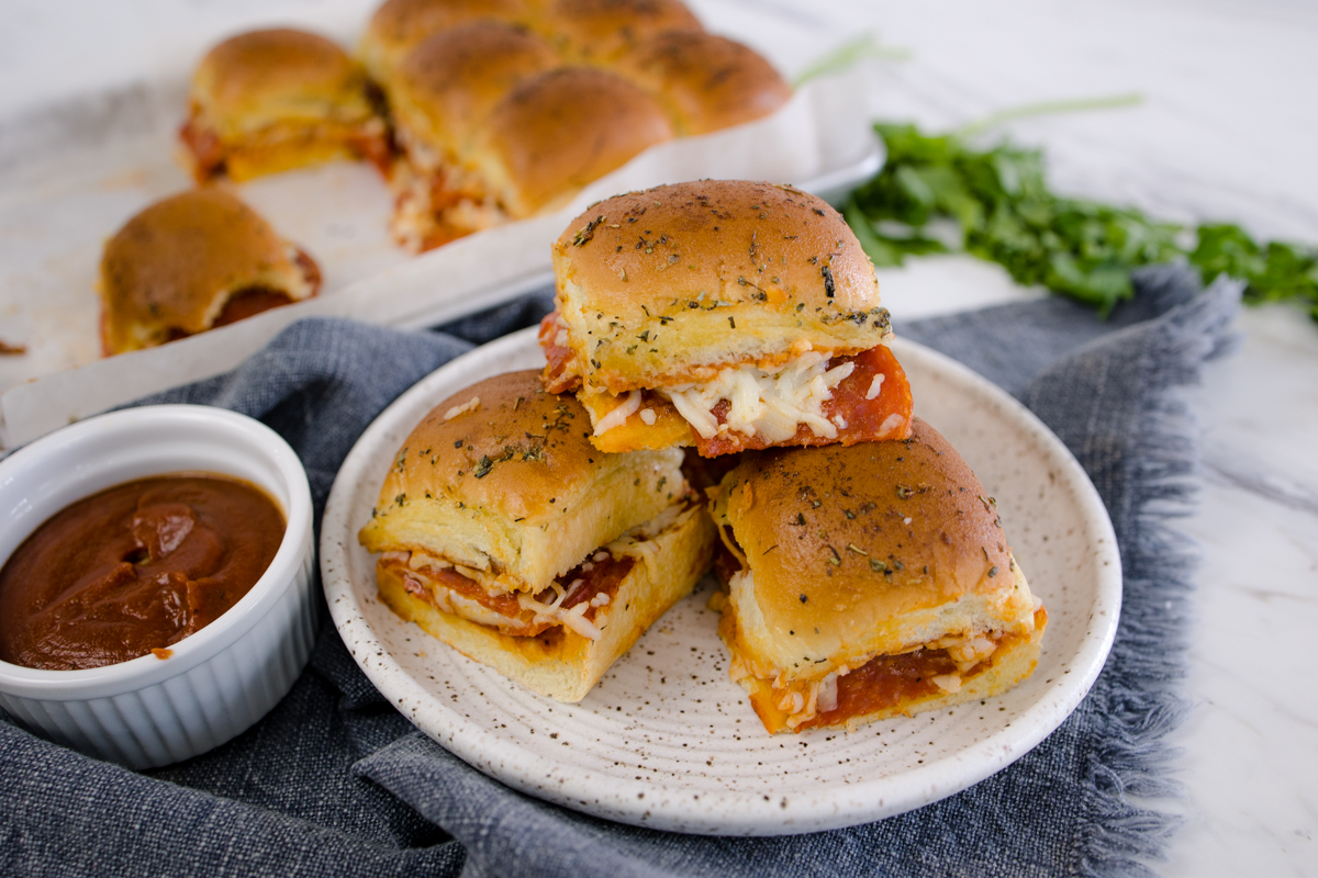 Top view of pizza sliders on a white plate, on a table with a small bowl of Marinara sauce on it and more pizza sliders on a baking tray in the background. 