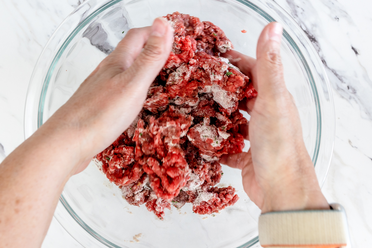 Top view of a glass mixing bowl with raw ground beef in it being mixed together with herbs by two hands. 