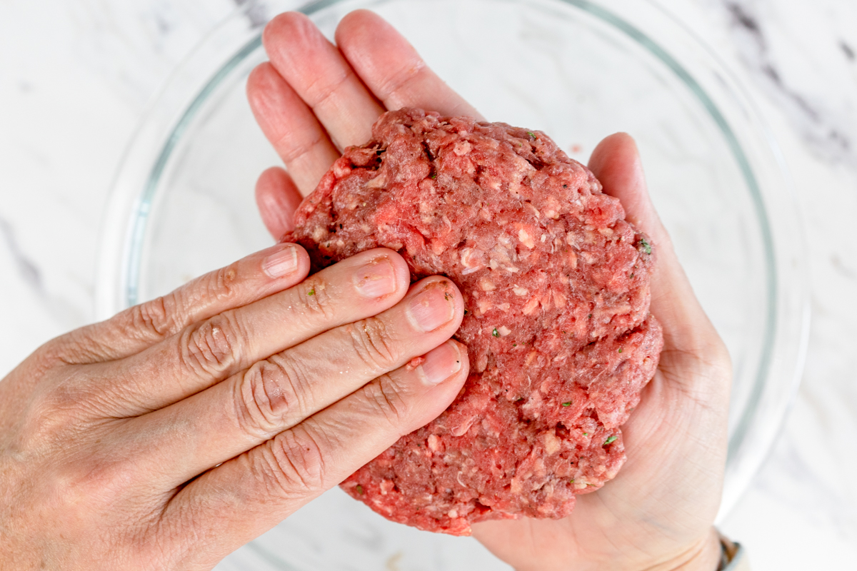 Top view of two hands shaping ground beef into patties in mid-air above a glass mixing bowl. 