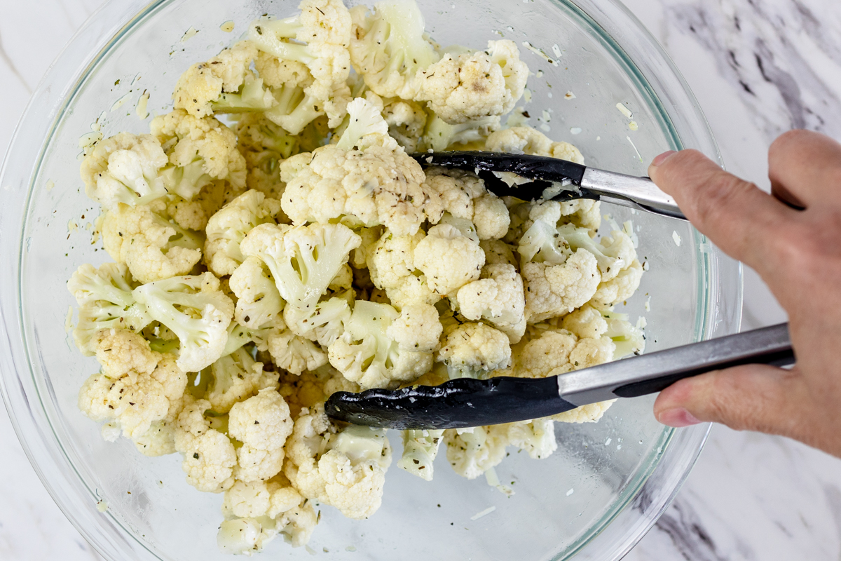 Top view of glass mixing bowl with cauliflower, seasoning, and cheese in it that is being tossed by someone holding tongs.