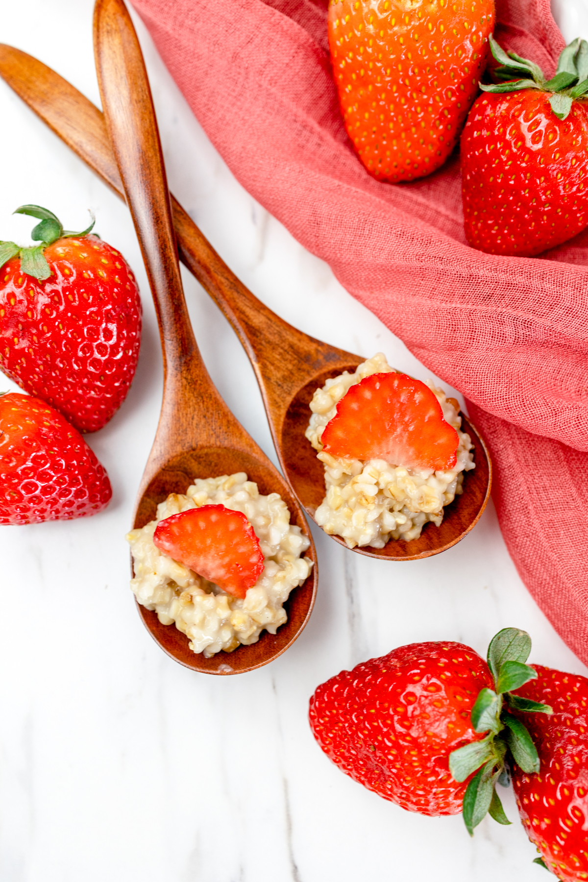 Top view of two wooden spoons with steel cut oat porridge in them and a slice of strawberry on top, on a table next to fresh strawberries. 