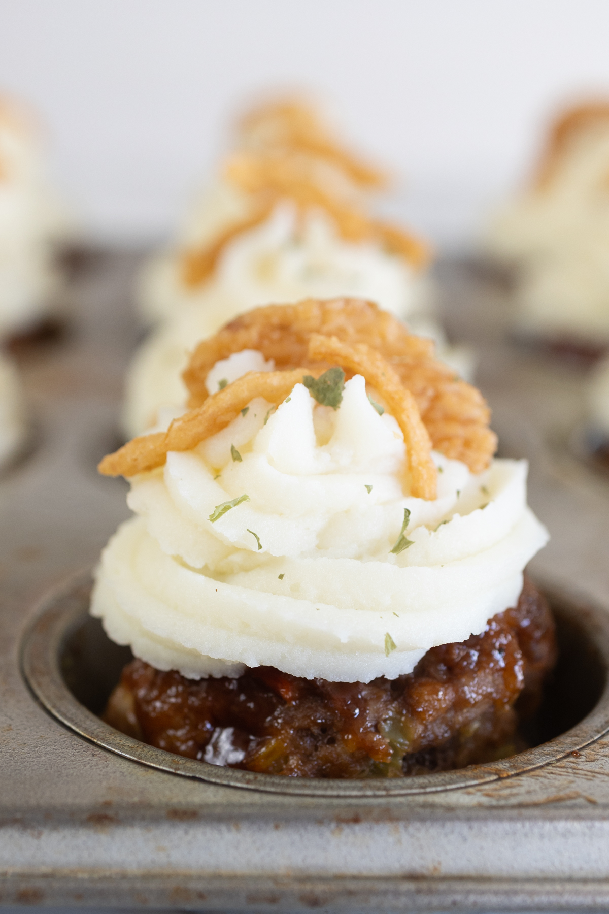 Close up of a meatloaf muffin in a muffin tin with mashed potatoes on top, garnished with French Fried Onions and some herbs. 