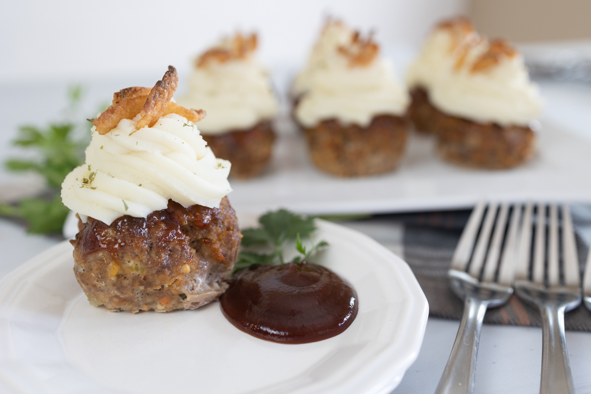 Close up of a meatloaf muffin on a white plate next to brown sauce and cutlery. 