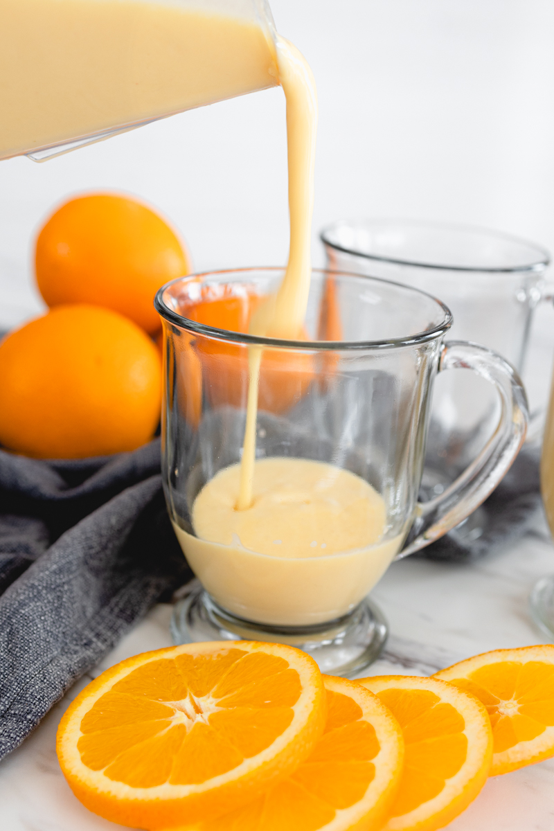 Orange Julius being poured into a glass mug, surrounded by slices of fresh orange.