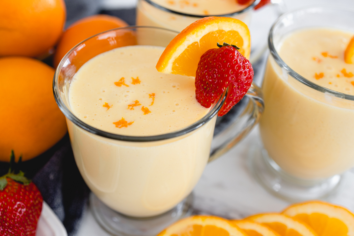 Close up of a glass mug of Orange Julius with a strawberry and a slice of orange on the side and orange zest on top.