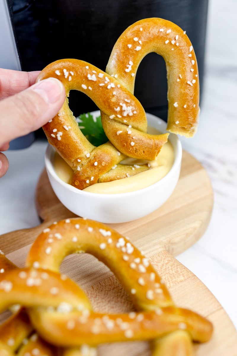 A hand holding an air-fried pretzel, dipping it in sauce, with an air fryer in the background.