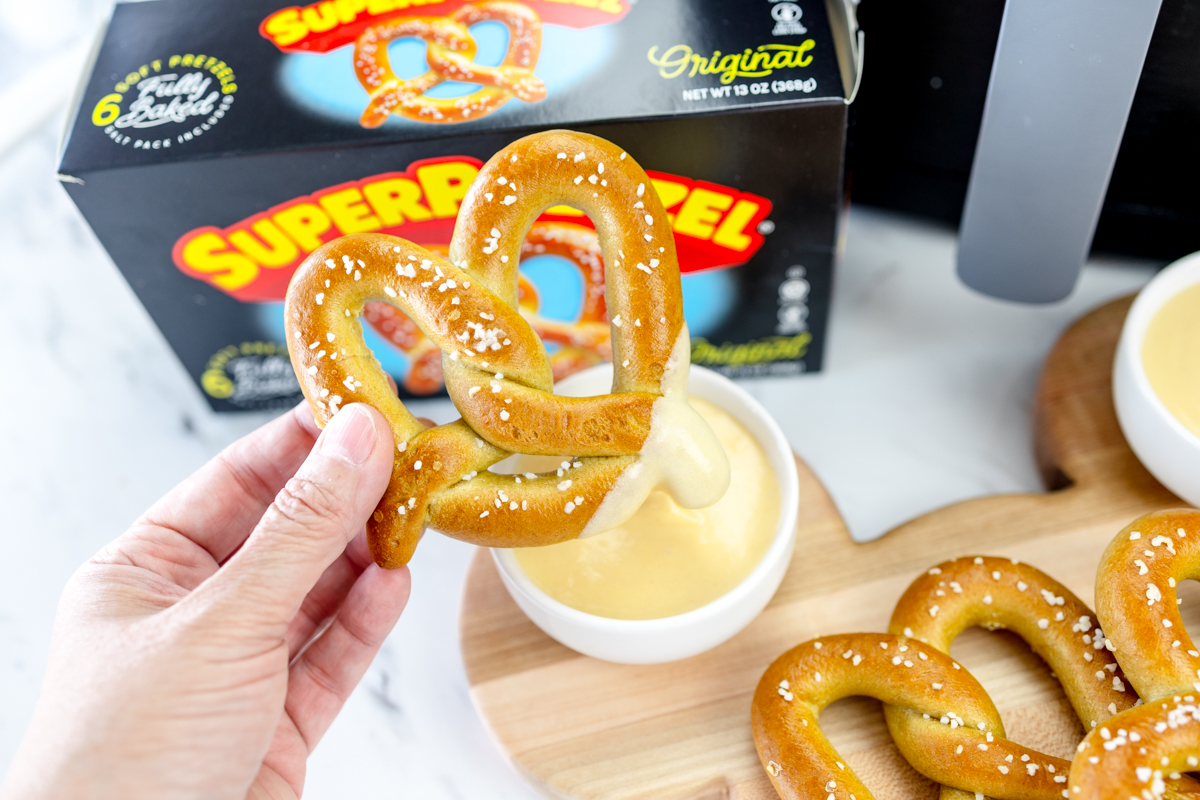A hand holding an air-fried pretzel which has been dipped in sauce, with a box of Super Pretzel brand frozen pretzels in the background.