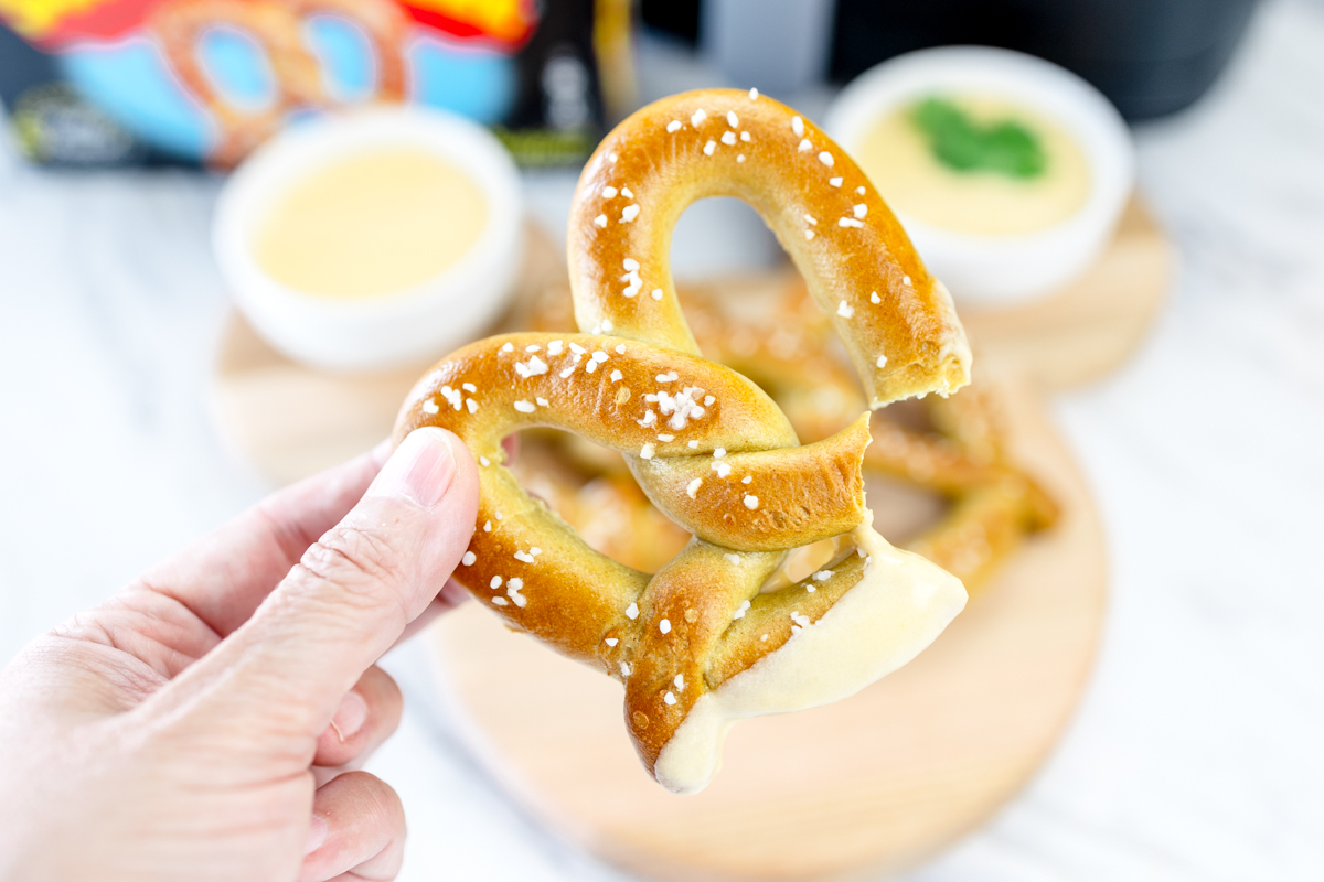 A hand holding an air-fried pretzel which has been dipped in sauce, with ramekins of sauce in the background.