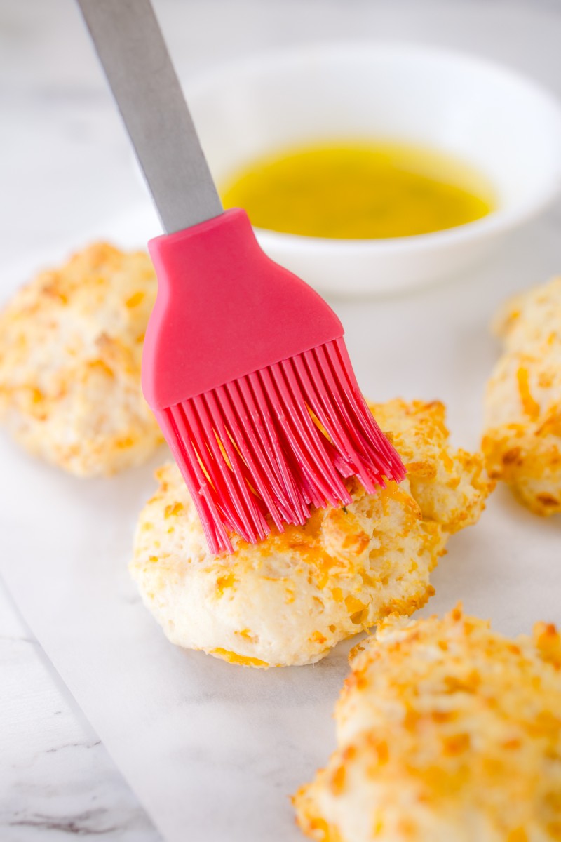 Close up of a silicone baster brushng seasoniing and melted butter over baked biscuits.