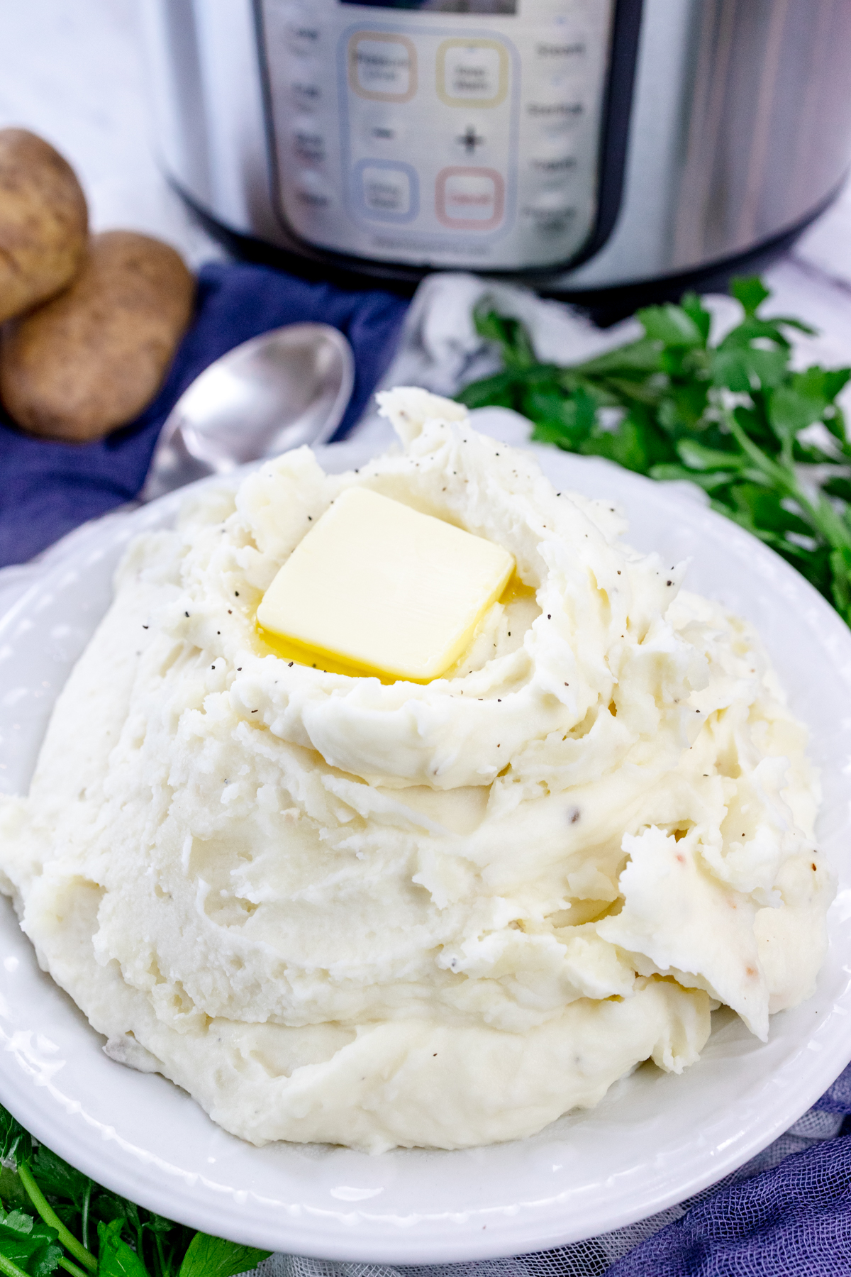 Top view of mashed potatoes with a melting chunk of butter on top and a spoon sticking out of it, in a white bowl on a table with an Instant Pot in the background, and decorations and ingredients around the bowl.