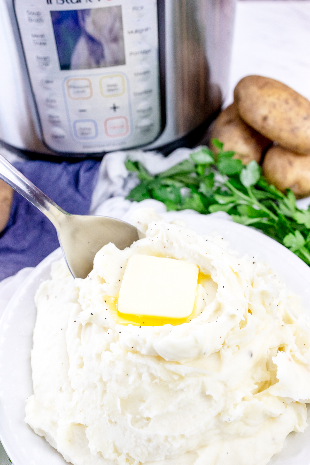 Top view of mashed potatoes with a melting chunk of butter on top and a spoon sticking out of it, in a white bowl on a table with an Instant Pot in the background, and decorations and ingredients around the bowl.