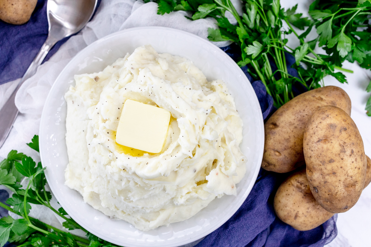Top view of mashed potatoes with a melting chunk of butter on top, in a white bowl on a table with decorations and ingredients around it.
