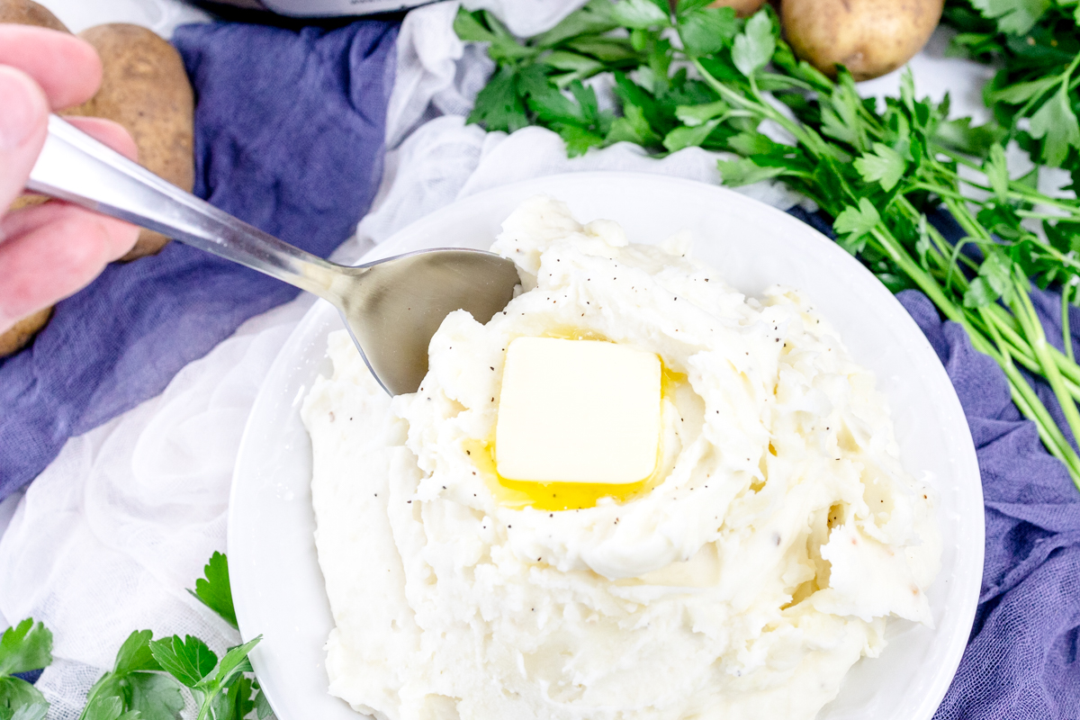 Top view of mashed potatoes with a melting chunk of butter on top and a spoon sticking out of it, in a white bowl on a table with decorations and ingredients around it.