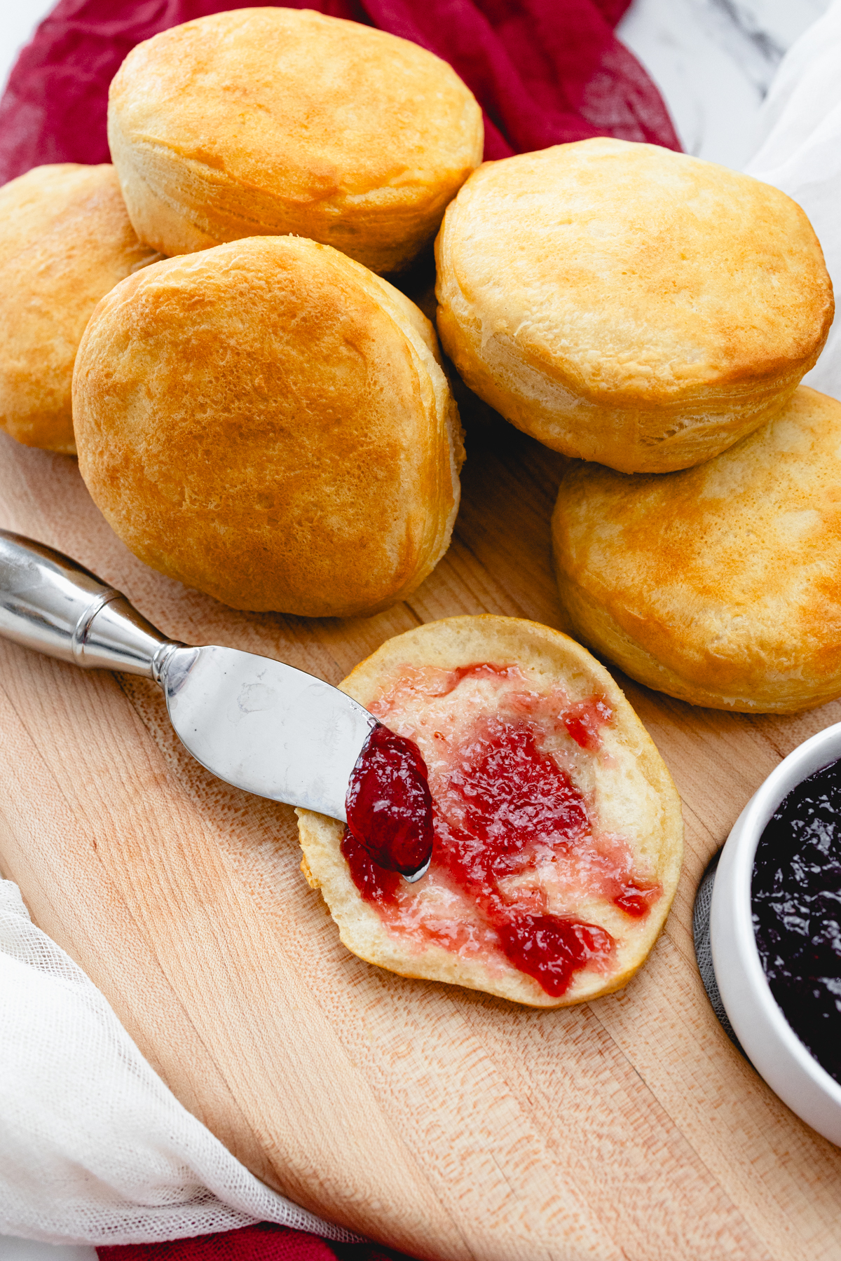 Top view of Pillsbury Grand Biscuits fresh out of the air fryer, on a wooden surface, with the one in front sliced open and a knife is spreading a berry jam onto it.