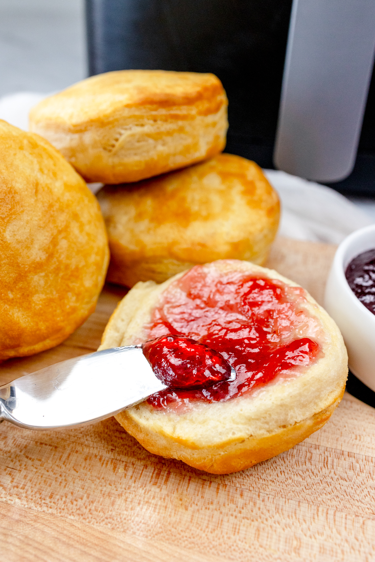 Close up view of Pillsbury Grand Biscuits fresh out of the air fryer, on a wooden surface, with the one in front sliced open and a knife is spreading a berry jam onto it.