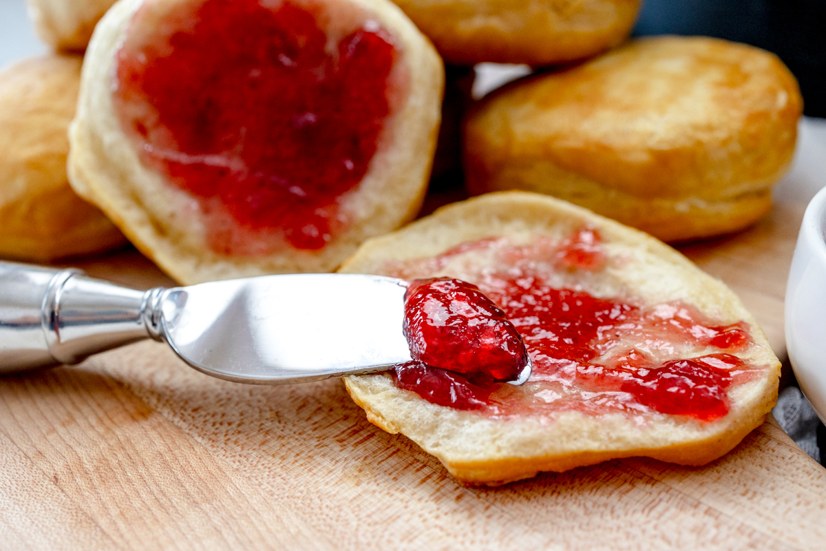 Close up view of Pillsbury Grand Biscuits fresh out of the air fryer, on a wooden surface, with the one in front sliced open and a knife is spreading a berry jam onto it.