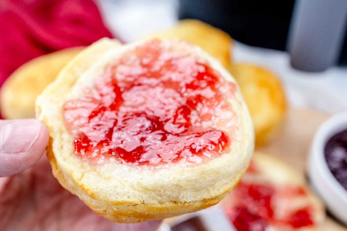 Close up view of a hand holding a Pillsbury Grand Biscuit fresh out of the air fryer, with berry jam spread on it.