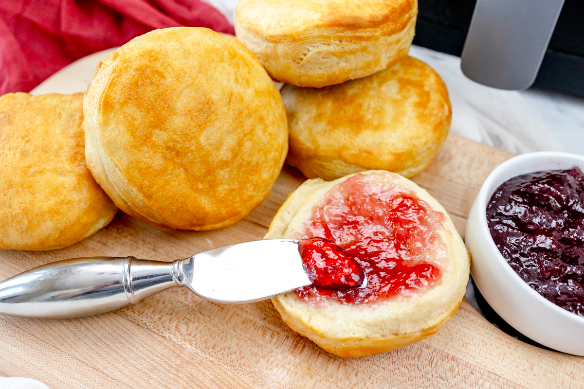 Close up view of Pillsbury Grand Biscuits fresh out of the air fryer, on a wooden surface, with the one in front sliced open and a knife is spreading a berry jam onto it.