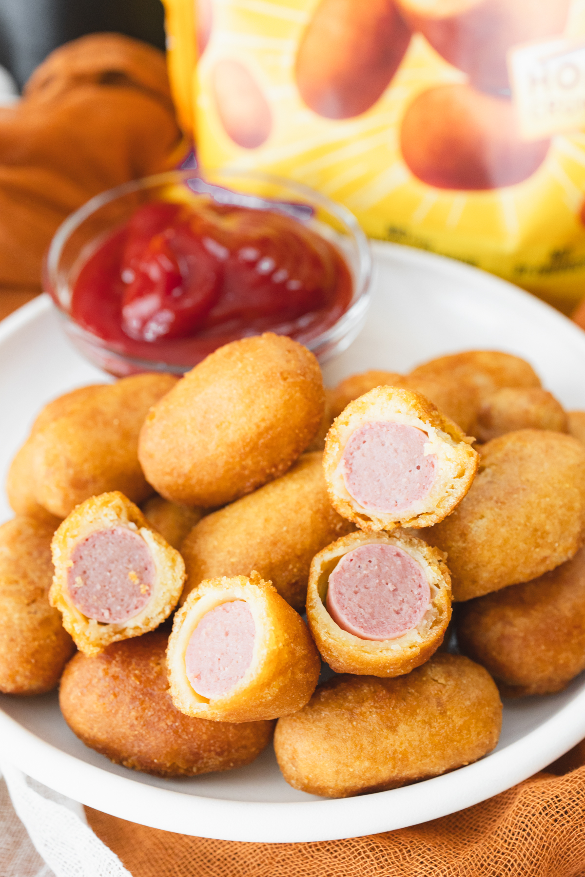 Close view of cooked mini corn dogs on a white plate that has a small dipping bowl of ketchup on it. IN the background is the bag of mini corn dogs.