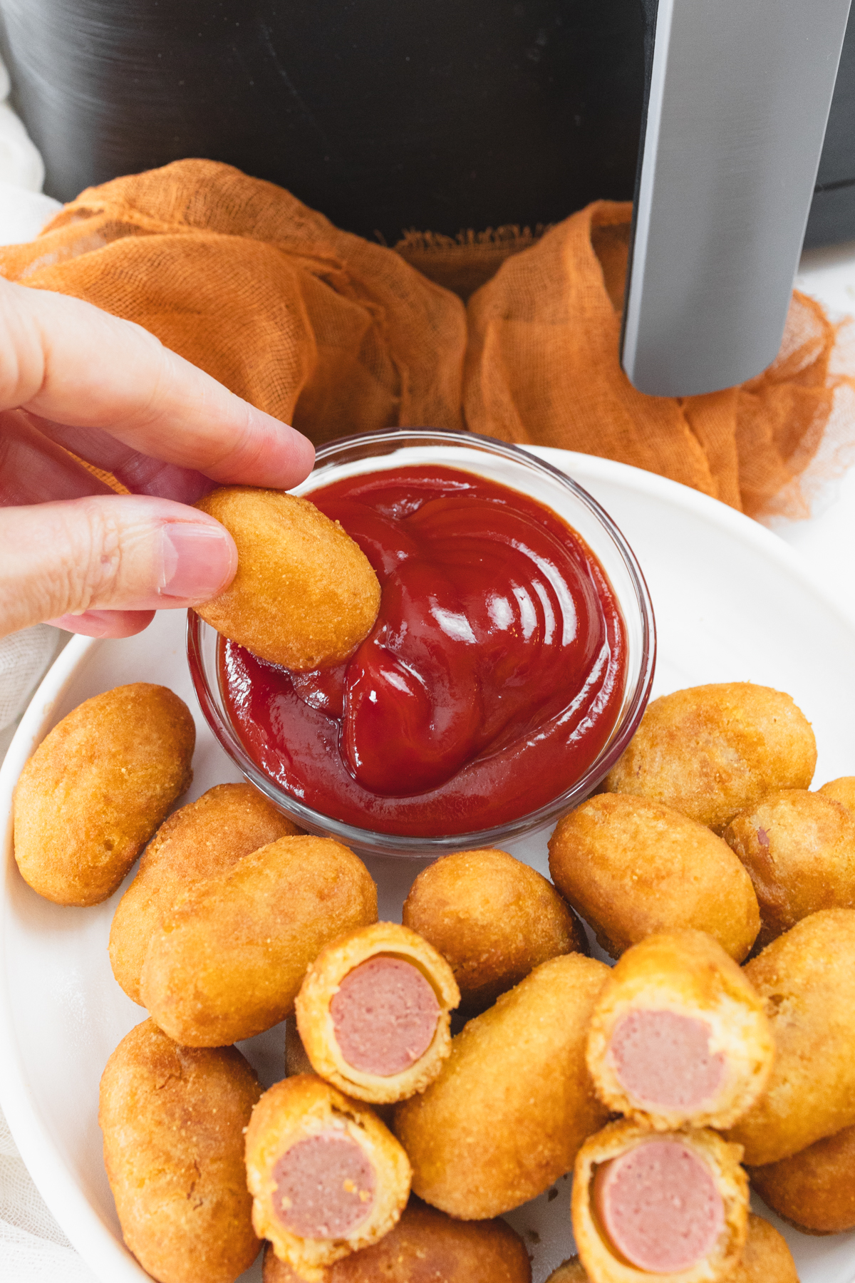 Close view of cooked mini corn dogs on a white plate that has a small dipping bowl of ketchup on it with a corn dog being dipped. IN the background is the air fryer.