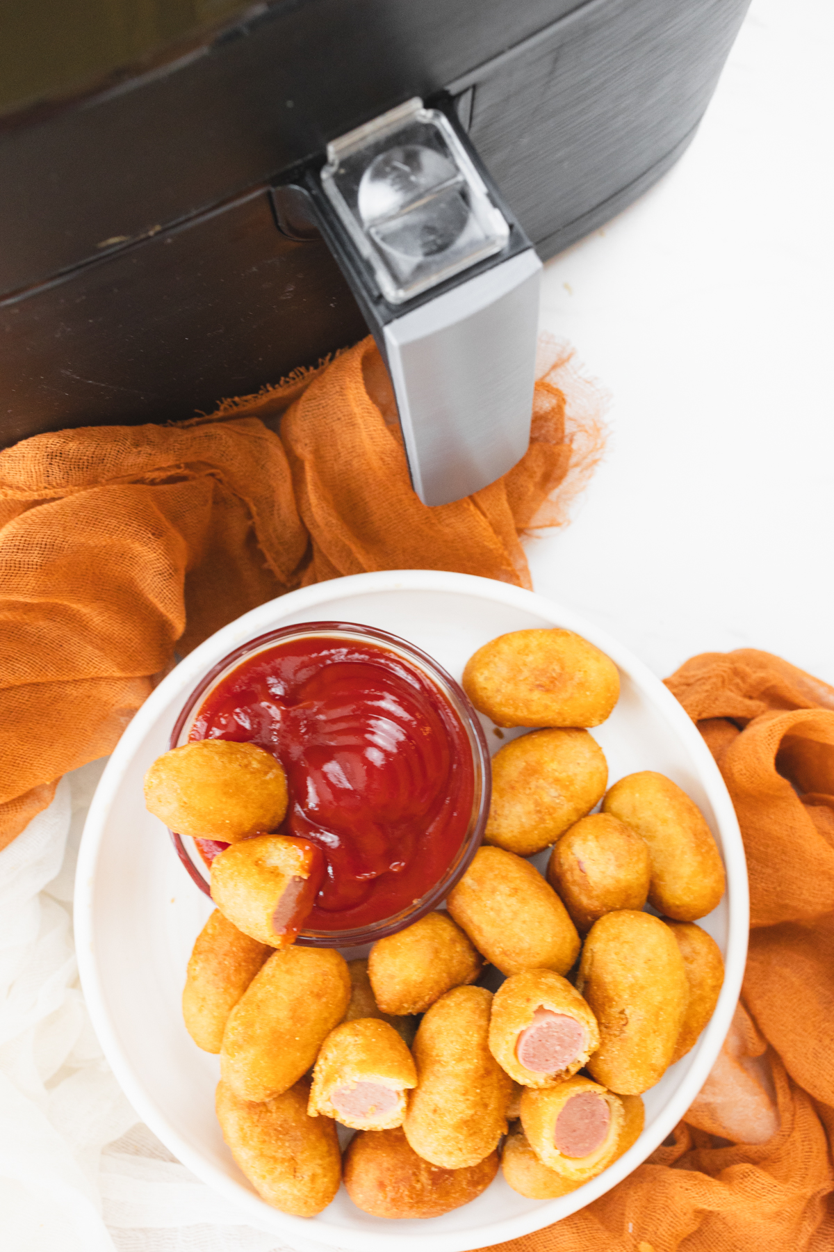 Close view of cooked mini corn dogs on a white plate that has a small dipping bowl of ketchup on it. In the background is the air fryer.