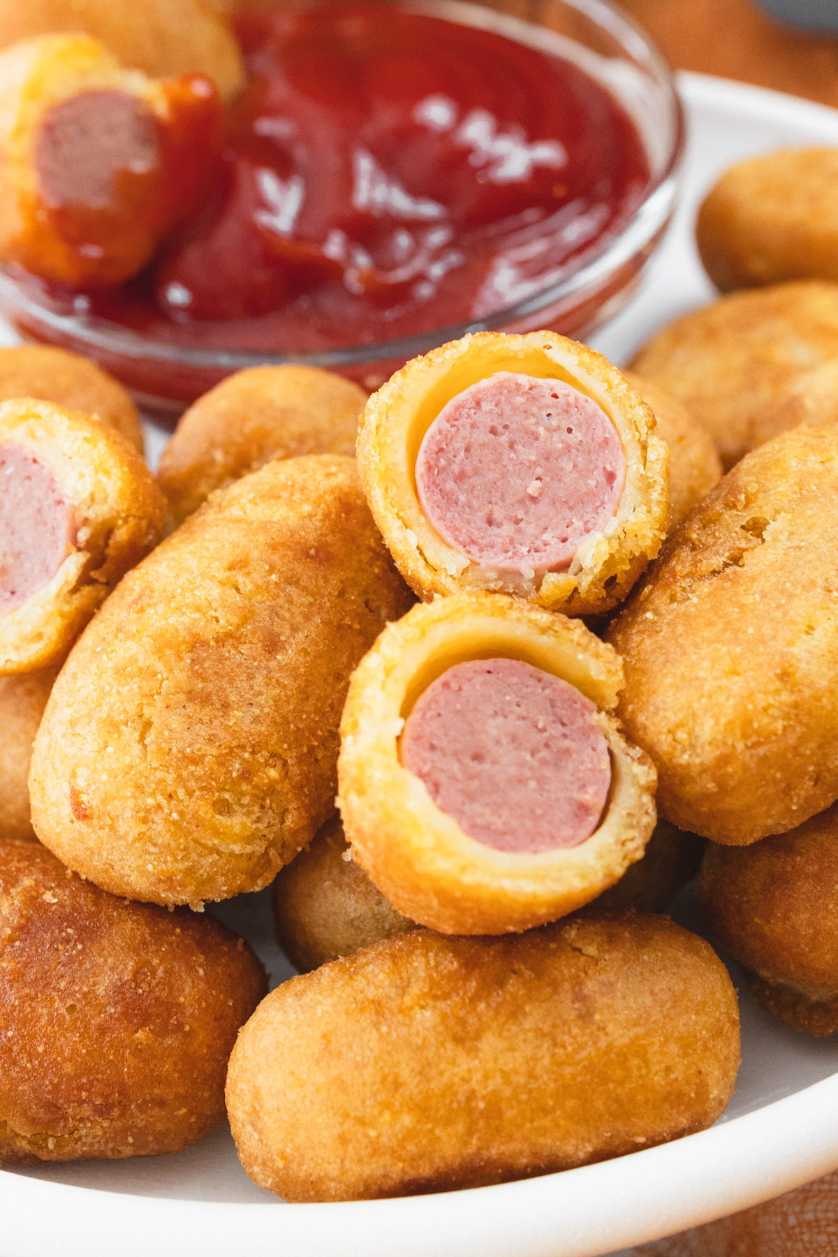 Close view of cooked mini corn dogs on a white plate that has a small dipping bowl of ketchup on it.