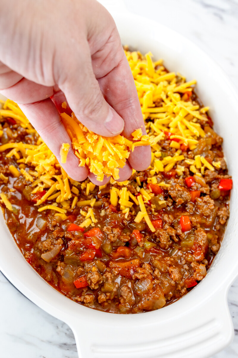 Top view of a hand sprinkling cheese over the sloppy joe mixture in the casserole dish.