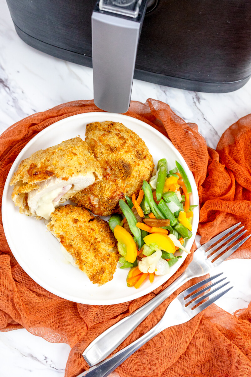 Overhead view of Air Fried Chicken Cordon Bleu on a white plate with side vegetables on the side, in front of an air fryer.