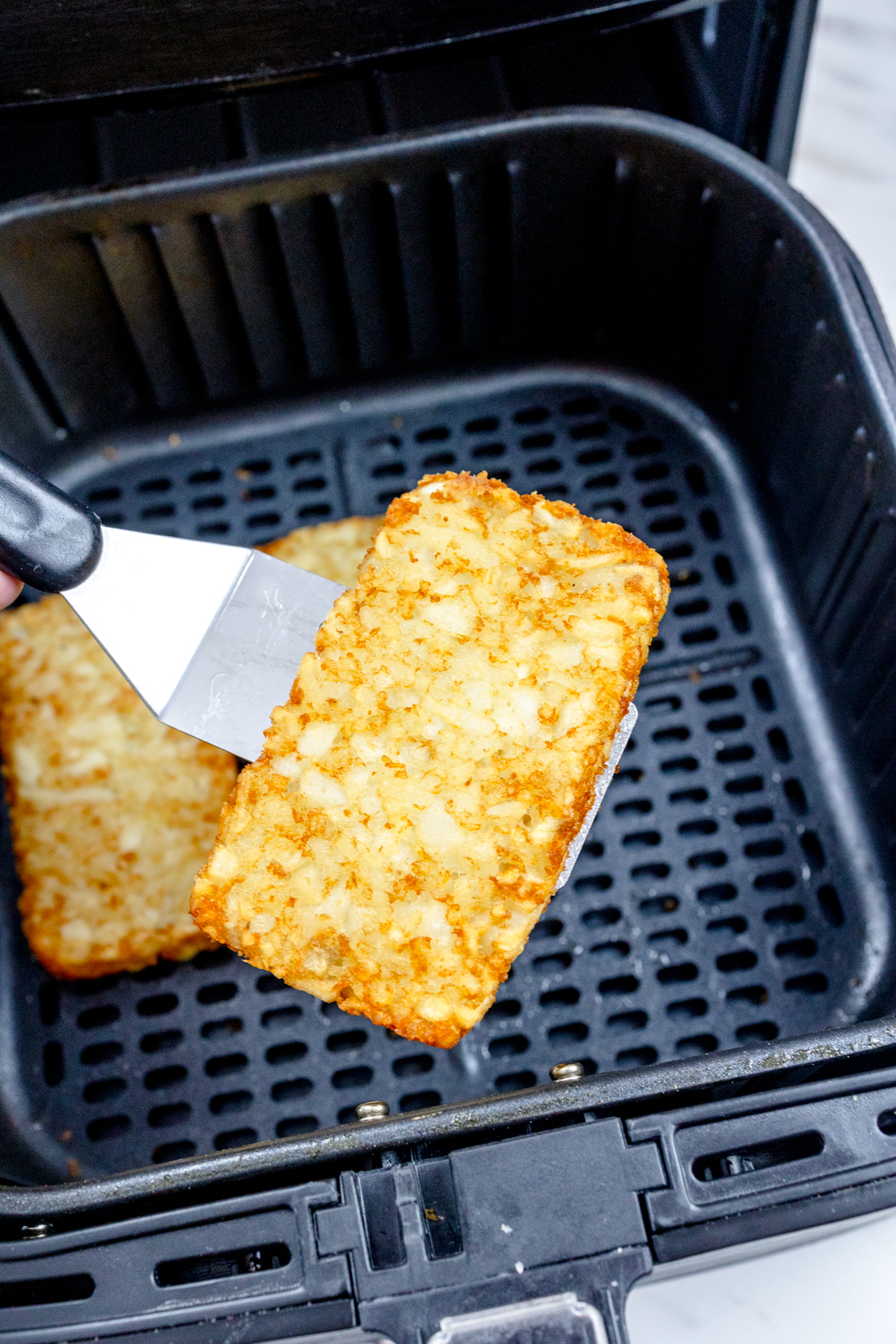 Close up view of a metal spatula lifting a hash brown out of an air fryer.