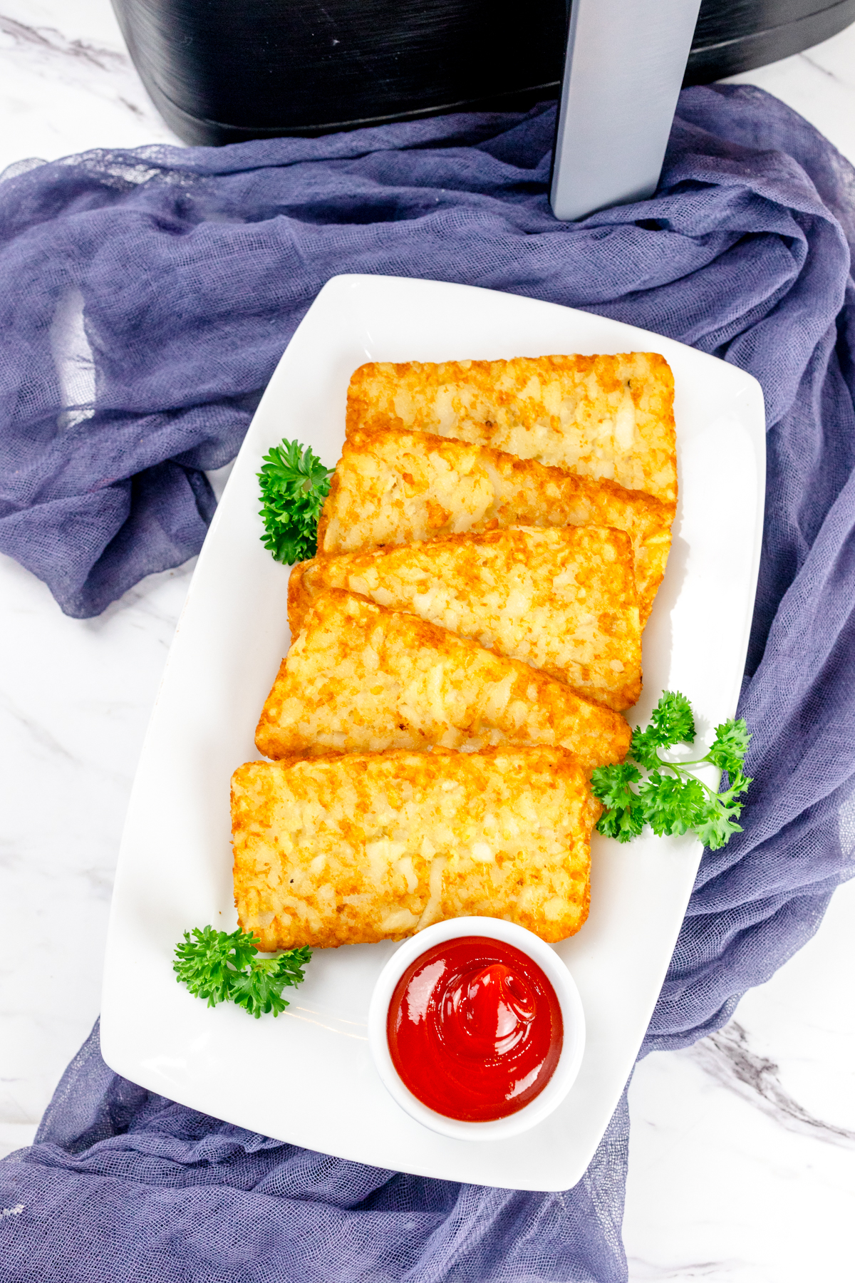 Top view of air fried hash browns on a white plate with leafy green garnish and a small dipping pot of tomato sauce.