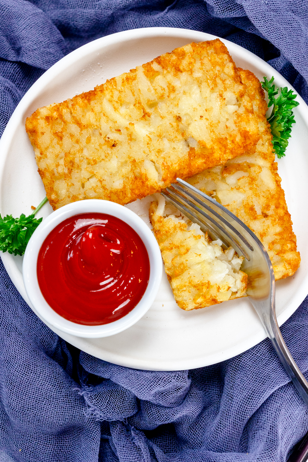 Top view of air fried hash browns on a white plate with leafy green garnish, a fork cutting one of the hash browns, and a small dipping pot of tomato sauce.