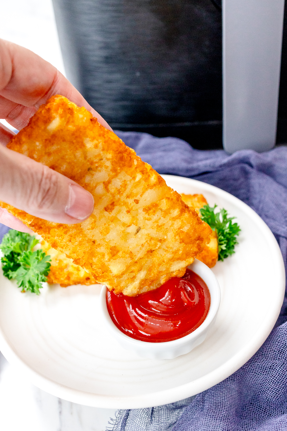 Close up view of a hand dipping a hash brown into a pot of tomato ketchup, on a white plate.