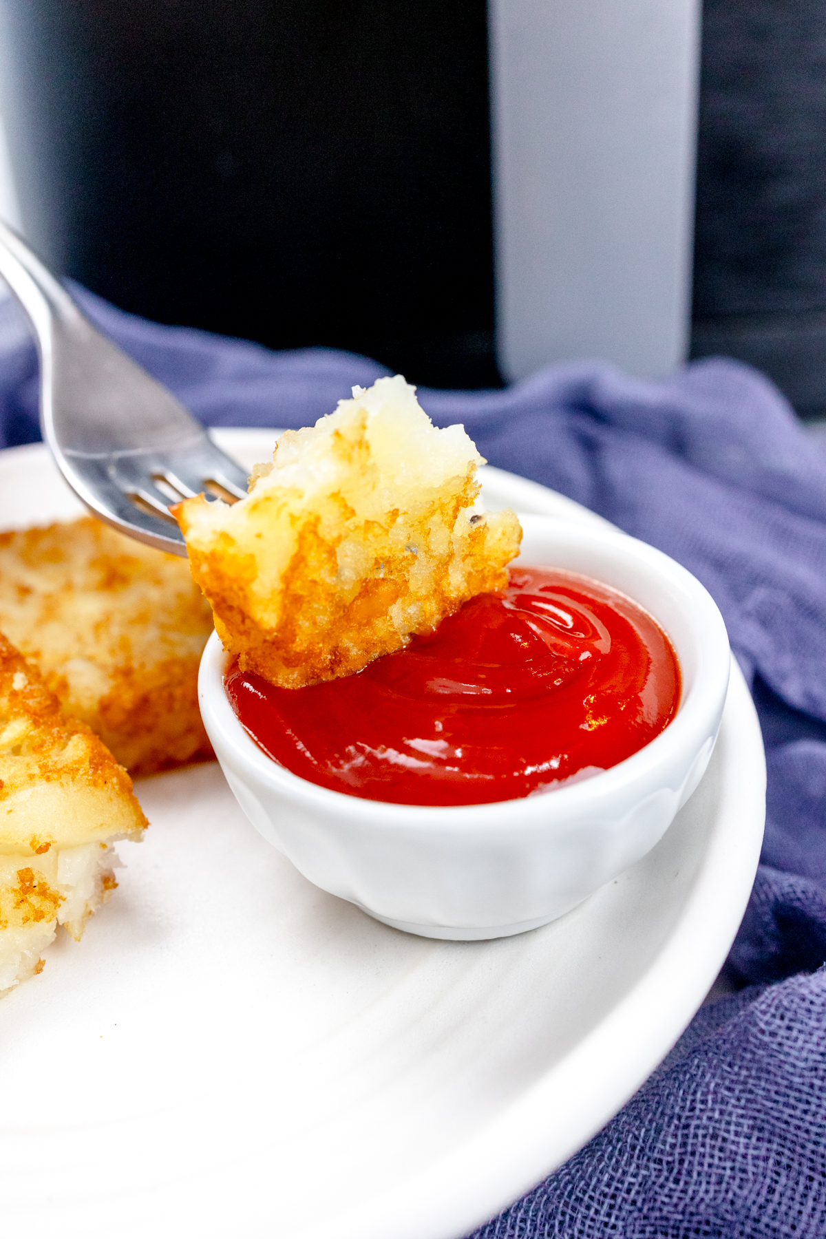 Close up view of a fork dipping a piece of hash brown into a pot of tomato ketchup, on a white plate.