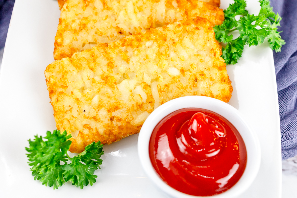 Top view of air fried hash browns on a white plate with leafy green garnish and a small dipping pot of tomato sauce.