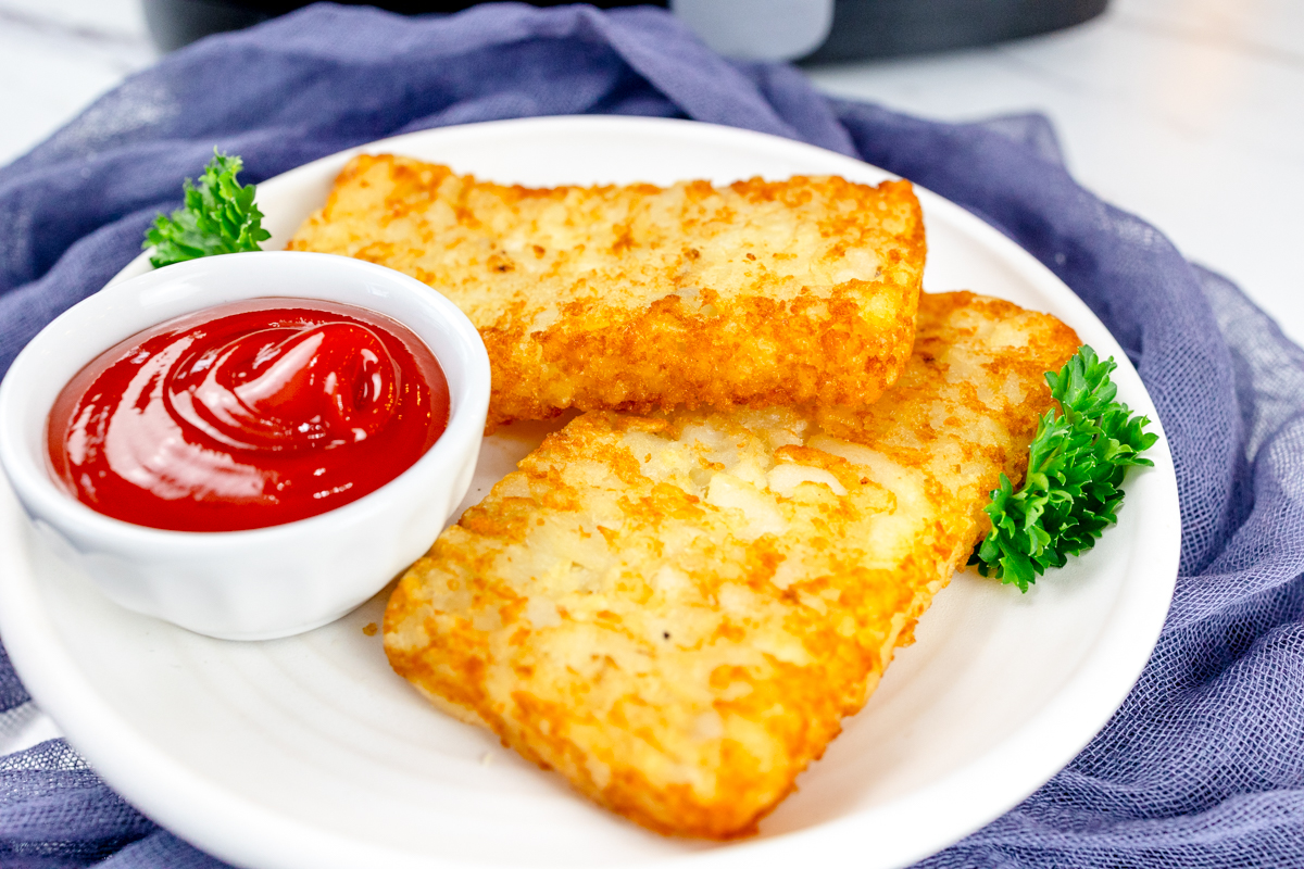 Close up view of air fried hash browns on a white plate with leafy green garnish and a small dipping pot of tomato sauce.