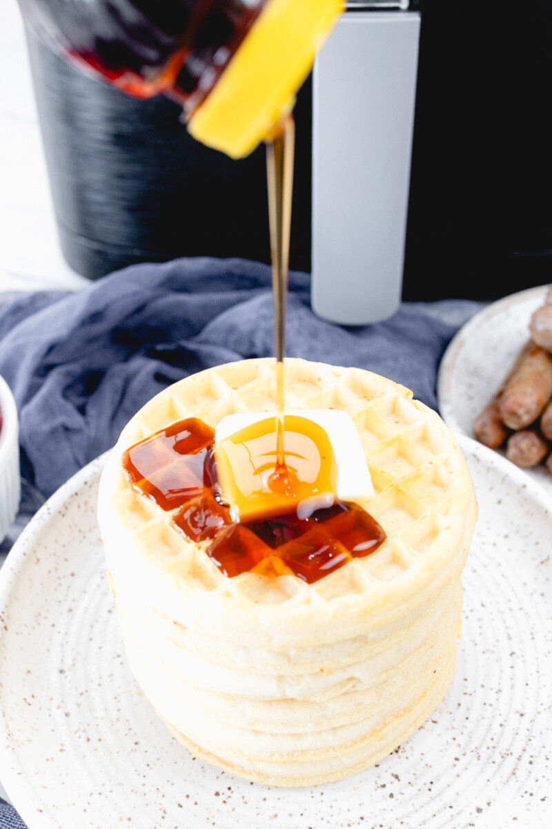 Close up view of syrup being poured onto a stack of waffles with some butter on top.